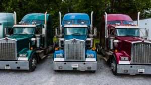Three semi truck cabs in green blue and maroon parked side by side facing the camera on a gravel lot