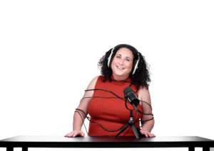 Smiling woman in a red top wearing over ear headphones and a headset mic at a table ready to record a podcast
