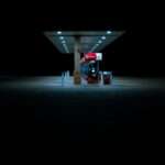 Gas station at night under a long lit canopy with multiple pumps and blue tinted lighting in the foreground