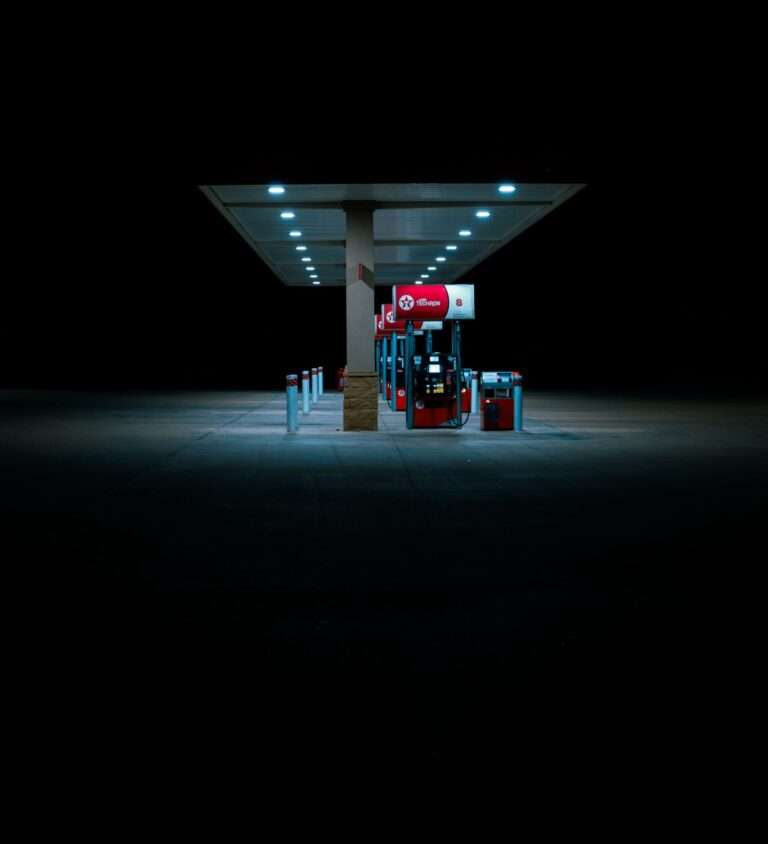 Gas station at night under a long lit canopy with multiple pumps and blue-tinted lighting in the foreground