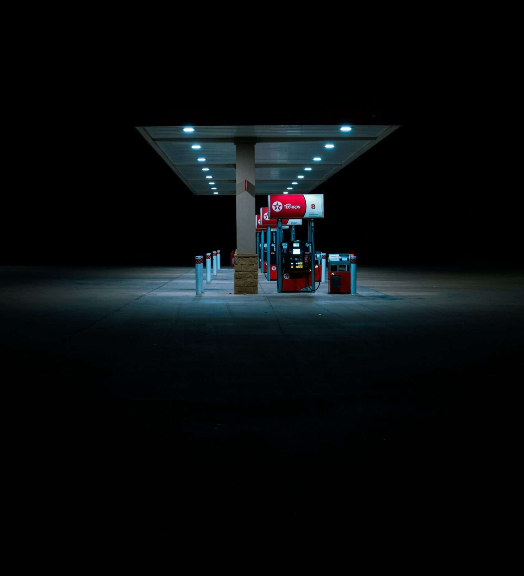 Gas station at night under a long lit canopy with multiple pumps and blue tinted lighting in the foreground