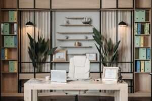 Modern executive office with a light wooden desk white chair laptop framed certificates and houseplants against a shelving wall