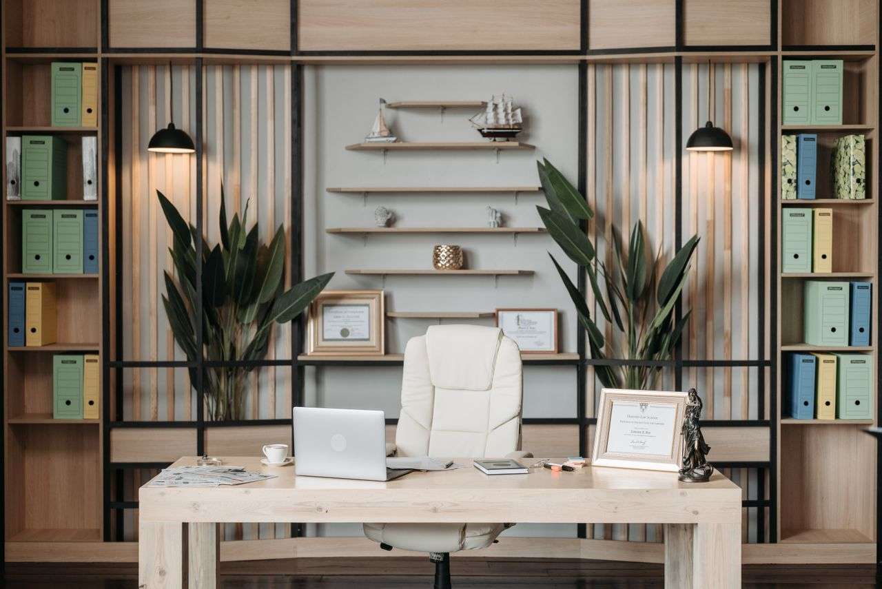 Modern executive office with a light wooden desk white chair laptop framed certificates and houseplants against a shelving wall