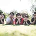 Six people lying on grass outdoors resting their chins on hands and making funny faces at the camera in a sunny park