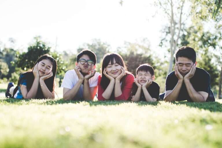 Six people lying on grass outdoors resting their chins on hands and making funny faces at the camera in a sunny park