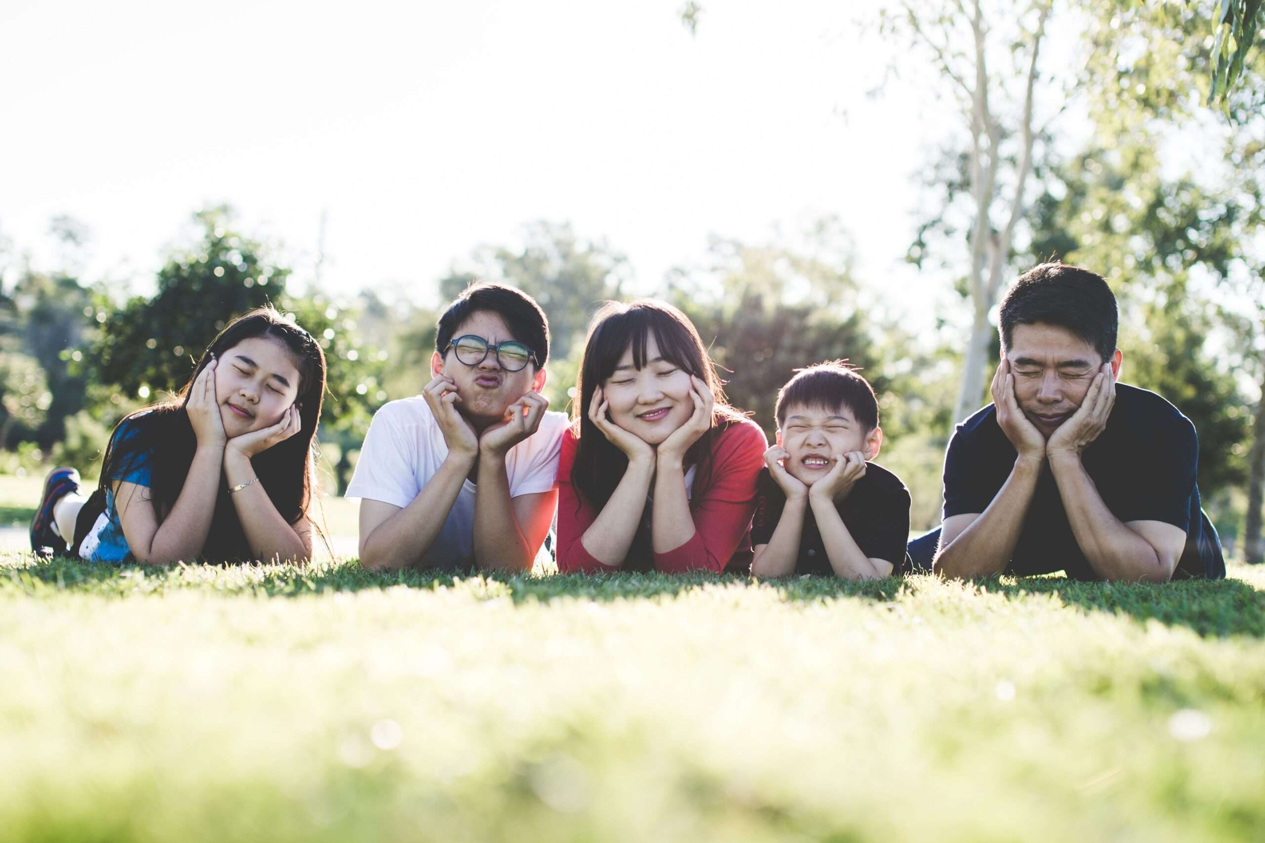 Six people lying on grass outdoors resting their chins on hands and making funny faces at the camera in a sunny park
