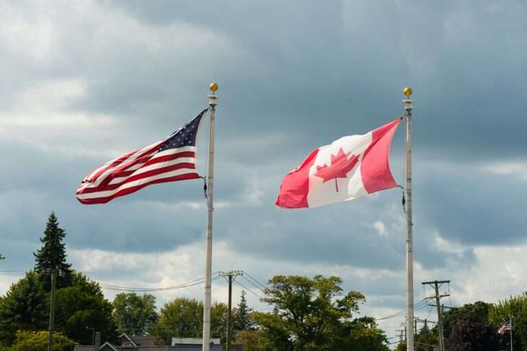 American and Canadian flags waving side by side on tall flagpoles against a cloudy sky.