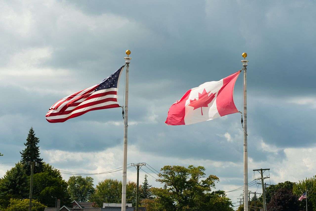 American and canadian flags waving side by side on tall flagpoles against a cloudy sky